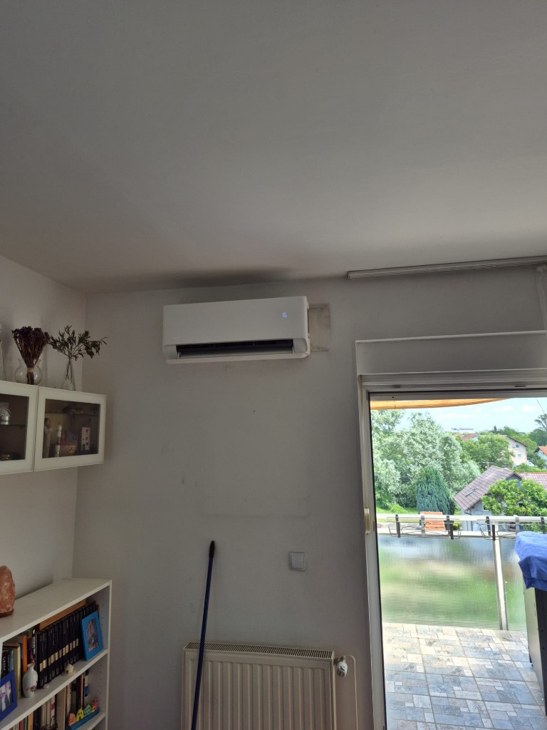Bijela klima na zidu dnevnog boravka, visoko iznad radijatora, pored balkonskih vrata.