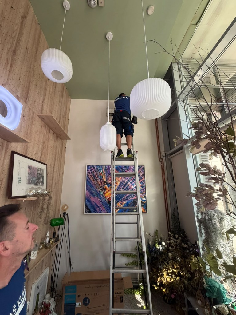 Monter na ljestvama postavlja unutarnju jedinicu u uređenom lokalu sa zelenim stropom i visećim svjetiljkama.
